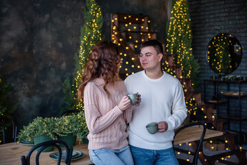 A loving couple in warm sweaters hold two turquoise ceramic cups in their hands, drink hot drinks in winter, against the background of Christmas lights. The atmosphere of winter holidays at home