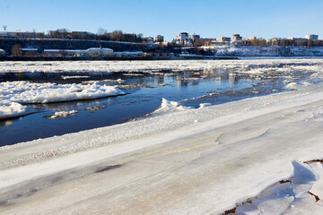 Ice drift on a river with blue high water and big water, white snow broken ice full of hummocks in it and city with houses at a background in sunny spring day.