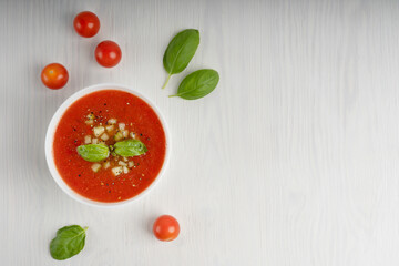 White bowl full of red gaspacho soup surrounded by whole tomatoes and green basil leaves standing on white wooden table. Image with copy space, horizontal orientation