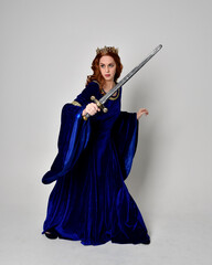 Full length portrait of  girl wearing long blue velvet gown with golden crown, holding a sword. standing pose  isolated against a studio background.