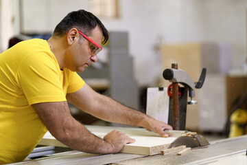 Carpenter cutting a piece of wood for furniture in his woodwork workshop, using a circular saw, and wearing safety goggles.