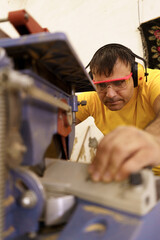 Carpenter cutting a piece of wood for furniture in his woodwork workshop, using a circular saw, and wearing safety goggles and earmuffs.