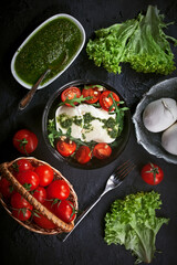 fresh mozzarella salad with lettuce and arugula leaves, pesto sauce, fresh cherry tomatoes and cedar nuts, on dark slate table background