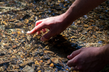 Male european hand refreshing in meditative zen atmosphere in a crystal clear water of a flat little creek with silky ripples and floating waves and sunny reflections searching for stones in the water