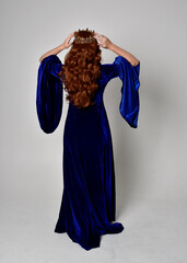 Full length portrait of  girl wearing long blue velvet gown with golden crown. standing pose with back to the camera, isolated against a studio background.