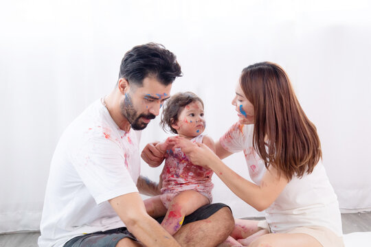Happy mixed race family and half-Turkish baby girl playing fun together paint face and paper, father, mother, parenthood hug adorable daughter paint color on dirty white clothes, hands and face