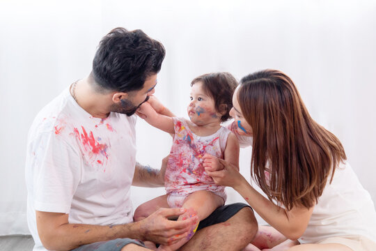 Happy mixed race family and half-Turkish baby girl playing fun together paint face and paper, father, mother, parenthood hug adorable daughter paint color on dirty white clothes, hands and face