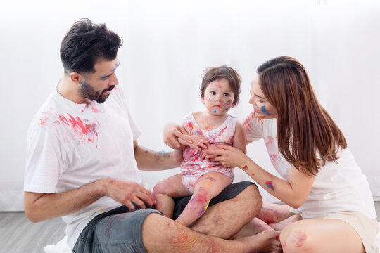 Happy mixed race family and half-Turkish baby girl playing fun together paint face and paper, father, mother, parenthood hug adorable daughter paint color on dirty white clothes, hands and face