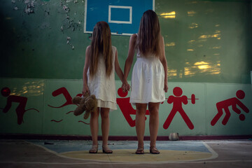 Two girls in white dresses with long hair stand in the abandoned gym of a destroyed school and hold...