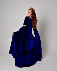 Naklejka premium Full length portrait of girl wearing long blue velvet gown with golden crown. standing pose with back to the camera, isolated against a studio background.