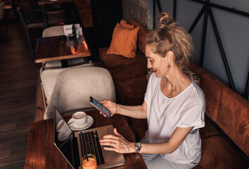 Smiling senior female blogger in casual wear sitting in cafe using laptop and smartphone.
