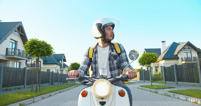 Portrait of attractive Caucasian delivery guy looking around for the customer house address. Young male courier riding on a scooter down to the street delivering the order. Self-isolation, quarantine.