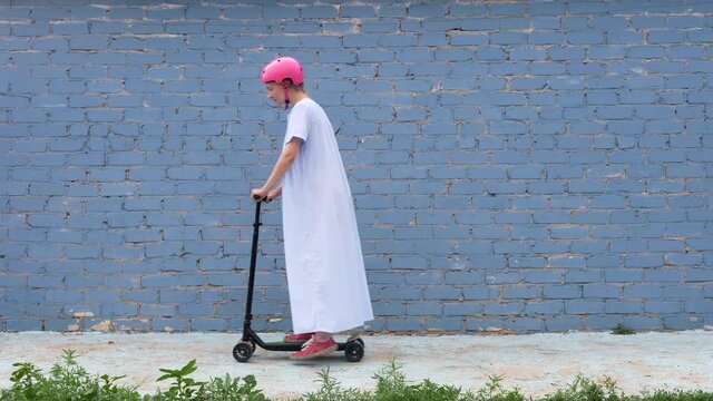 girl in pink helmet and long white dress with blond hair braided in pigtail is rolling on scooter against background of blue brick wall. blonde woman helmet for rolling kick scooter