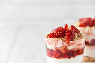 Homemade layered dessert with fresh strawberries, cream cheese or yogurt, granola and strawberry jam in glasses on white wood background. Healthy organic breakfast or snack concept. Selective focus.
