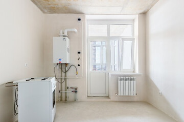 A small undecorated kitchen room with the gas-stove and the wall mounted water heater. The fine finished room with the white plastered walls in a new residential building