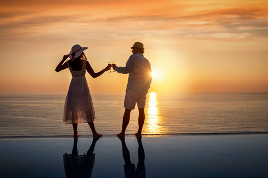 Ein glückiches Paar im Sommerurlaub steht am Randes eines Swimming Pools und genießt den Sonnenuntergang über dem Meer bei einem Glas Wein