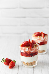 Homemade layered dessert with fresh strawberries, cream cheese or yogurt, granola and strawberry jam in glasses on white wood background. Healthy organic breakfast or snack concept. Selective focus.