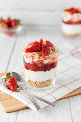 Homemade layered dessert with fresh strawberries, cream cheese or yogurt, granola and strawberry jam in glasses on white wood background. Healthy organic breakfast or snack concept. Selective focus.