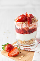 Homemade layered dessert with fresh strawberries, cream cheese or yogurt, granola and strawberry jam in glass on white wood background. Healthy organic breakfast or snack concept. Selective focus.