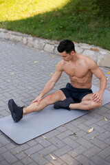 Obraz premium Young athlete with bare torso in sportswear doing stretching exercises on the mattress for sports in the park.