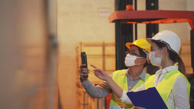 Asian Female Worker Talking With Manager In Warehouse Store. Woman And Man Engineers People Wear Safety Hard Helmet, Vest And Face Mask Checking Storage Box Parcel In Factory During Covid 19 Pandemic.