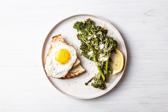 Roasted broccolini, lemon, fried egg with tost on the plate on the table. Top view on healthy food.