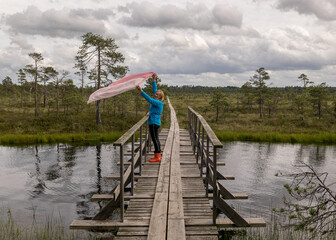 the summer swamp. woman in a blue jacket on a wooden bridge. bog pond. bog background and vegetation. white clouds. small swamp pines