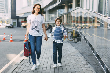 The mother carries a backpack in one hand, and holds her son by the other hand. A cheerful smiling boy walks with his mother from school