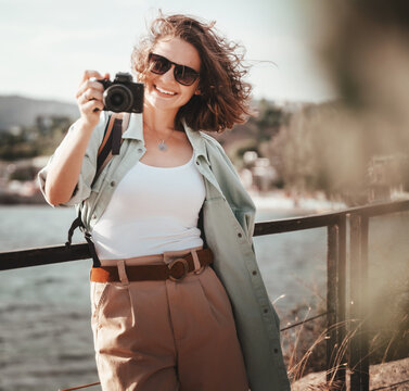 Beautiful curly trendy young hipster female at the sea in sunglasses with a photo camera in hand, summer vacation, travel, image retro film toning