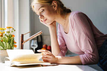 Photo of focused nice woman reading book while standing in kitchen