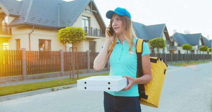Beautiful Caucasian courier with a backpack delivering food in cardboard boxes. Cheerful female delivery worker can't find the adress, calls and talks to customer looking for house. Delivery service.