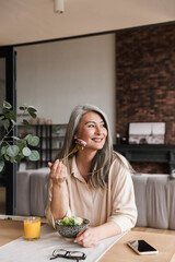 Woman sitting at kitchen indoors at home while eating salad.