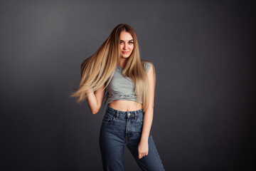 A thin girl with blond long hair looks away and straightens her hair against a dark wall