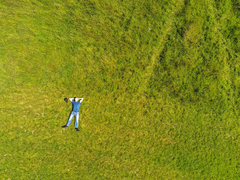 Man Lying On The Ground In A Park On A Green Grass Field. Man Dressed In Blue T Shirt And Jeans Hands Behind Hes Head. Aerial Top Down View.