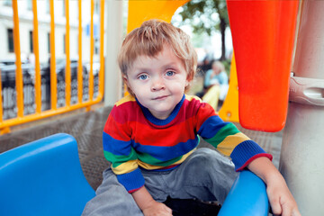 Fototapeta na wymiar A cool cheerful boy sits in the playground and smiles. A boy with a smile. The child is sitting on a children's slide. Baby in bright clothes in the playground. Mischievous boy with a rash smile