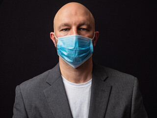 Portrait of a bald businessman in white t-shirt and grey suit on dark background, wearing blue protective face mark during COVID 19 flue outbreak.