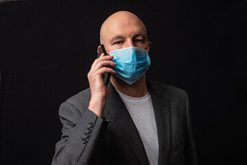 Portrait of a bald businessman in white t-shirt and grey suit on dark background, talking on black smart phone and wearing blue protective face mark during COVID 19 flue outbreak.