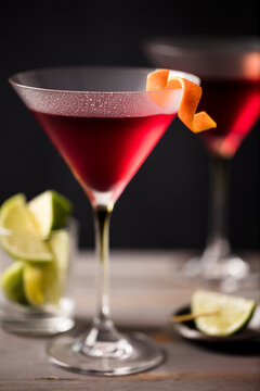Closeup 2 glasses of cosmopolitan cocktail decorated with orange served with lime wedges.  Dark grey worktop with dark background