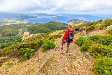 Hiking to Volterraio Castle. Backpacker woman looking views of Portoferraio Gulf, Elba Island. Stone stairs along Monte Volterraio with medieval fortress dominates.Tourism in Tuscany Italy destination - obrazy, fototapety, plakaty
