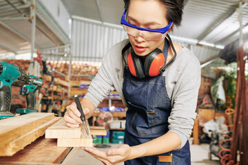Professional female carpenter in protective glasses drawing marks on wooden board before cutting it