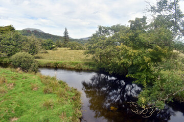 Fototapeta na wymiar Rustic scene in the Welsh countryside near to the Snowdonia National Park. A river meanders gently through the fields on a summers day. Trees line the bank, reflecting on the still waters.