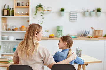 Back view portrait of caring mother hugging little girl while sitting at table and helping her study at home, copy space
