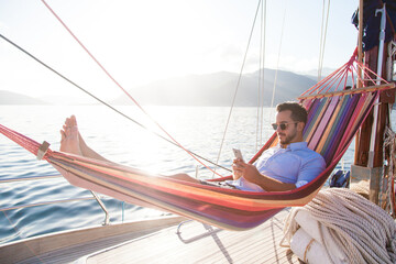 Man relaxing on yacht in hammock. Traveler with mobile phone. Travel on sailboat. Summer vacation...