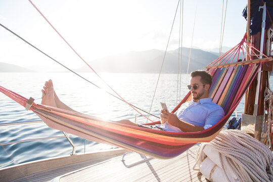 Man relaxing on yacht in hammock. Traveler with mobile phone. Travel on sailboat. Summer vacation in self isolation, social distance. Freelancer workplace in quarantine. Successful business lifestyle.