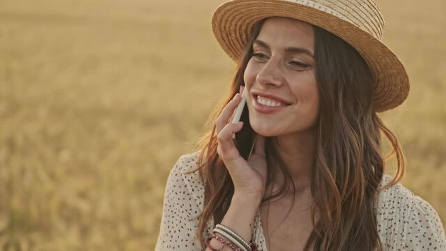 Happy attractive woman wearing dress and stylish hat talking by smartphone and laughing while walking on wheat field