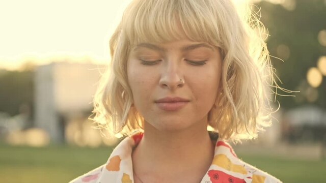 A pretty young blonde woman is opening her eyes and looking the camera while walking in the park
