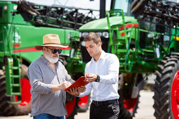 Fototapeta na wymiar Agreement. Professional farmer with a modern combine in sunlight at work. Confident, bright summer colors. Agriculture, exhibition, machinery, plant production. Senior man near his tractor with