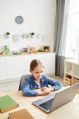 Vertical wide angle portrait of cute little girl using laptop and smiling while studying online at home in cozy interior , copy space