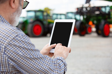 Professional farmer with a modern tractor at work with tablet. Looks confident, bright summer colors, sunshine. Agriculture, exhibition, machinery, plant production. Senior man near his machine.