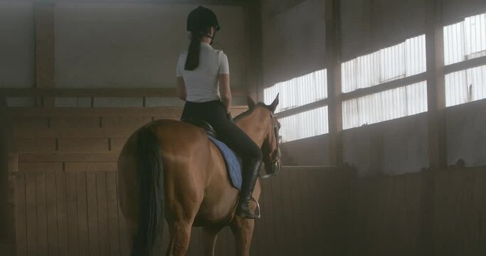 Young woman gets on a horse and riding a horse in a covered hangar, female trains riding on a horse, 4k slow motion.